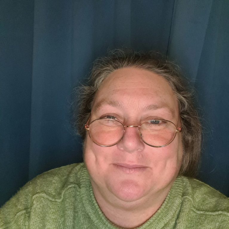 Smiling woman with glasses, wearing a green top, against a blue background.